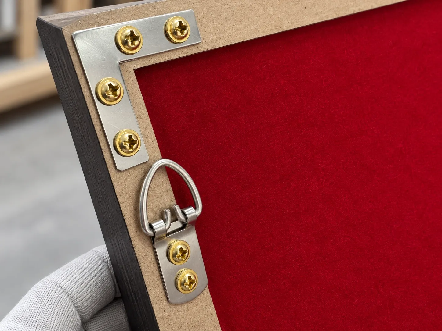 The back corner of a framed piece: a metal L-shaped bracket screwed across the joint and a D-ring hanger screwed in beside it, with a red fabric back behind.