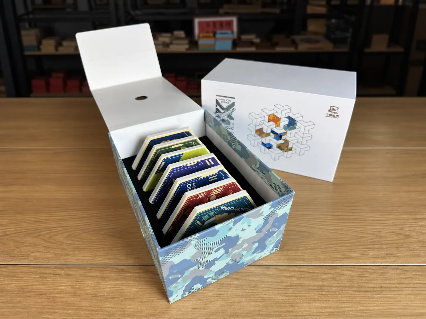 An open camouflage-printed flip-top box holding a series of shaped pieces in a foam insert, beside a printed white outer box.
