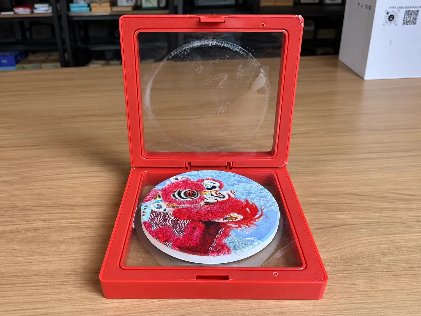 An open red floating display box with a round lion-dance disc held between two clear films.