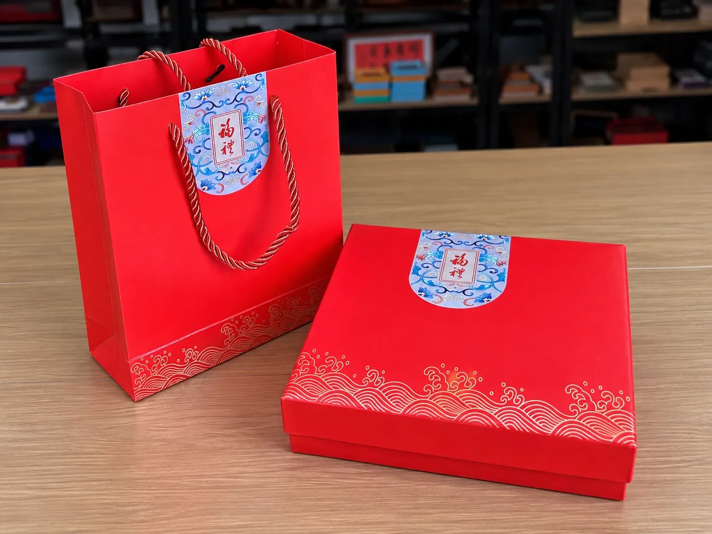 A red gift box and a matching red carrier bag with a gold wave border and a blue-and-white label.