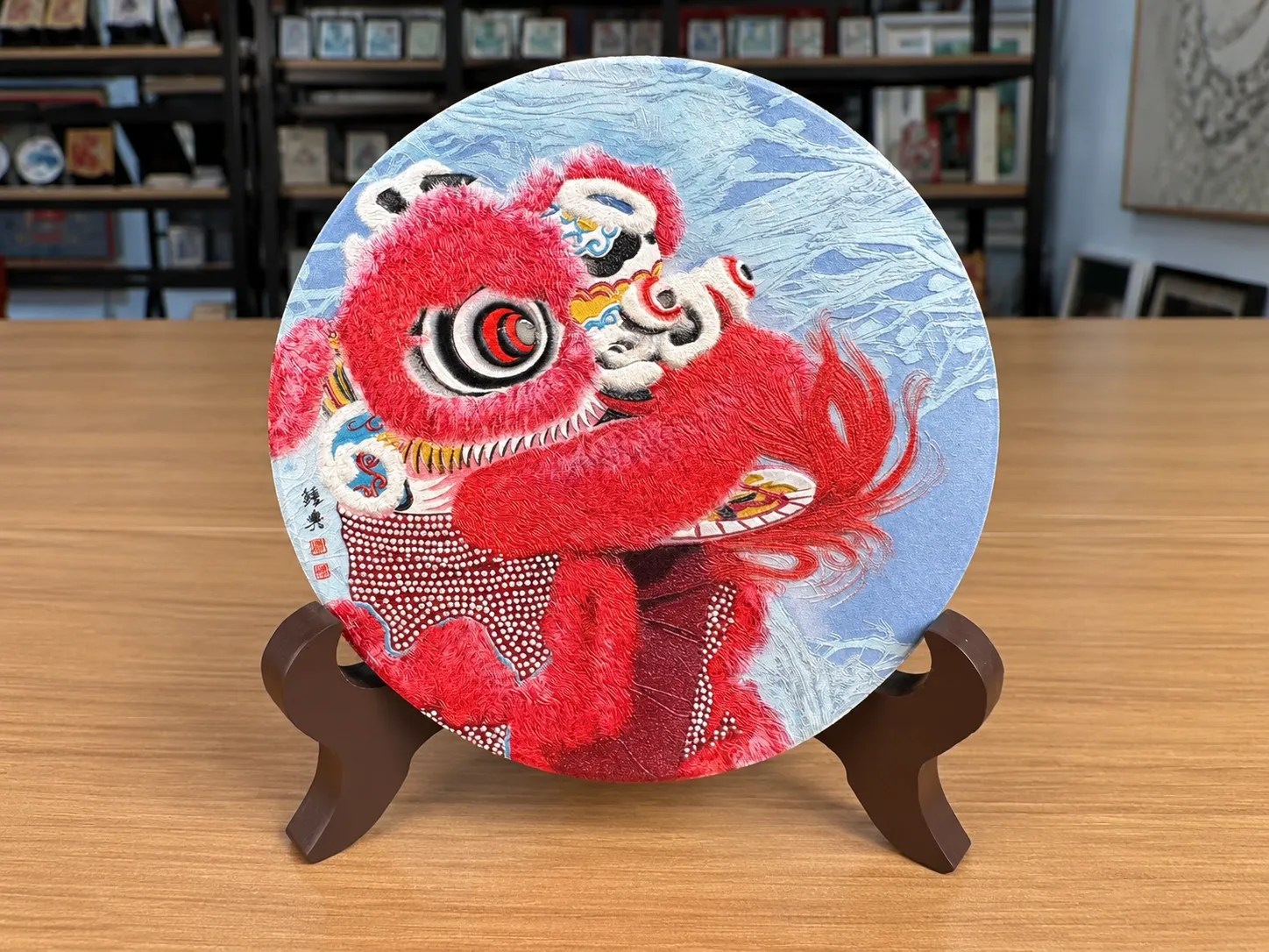 A round ceramic disc with a red lion-dance design standing on a dark wooden plate stand.