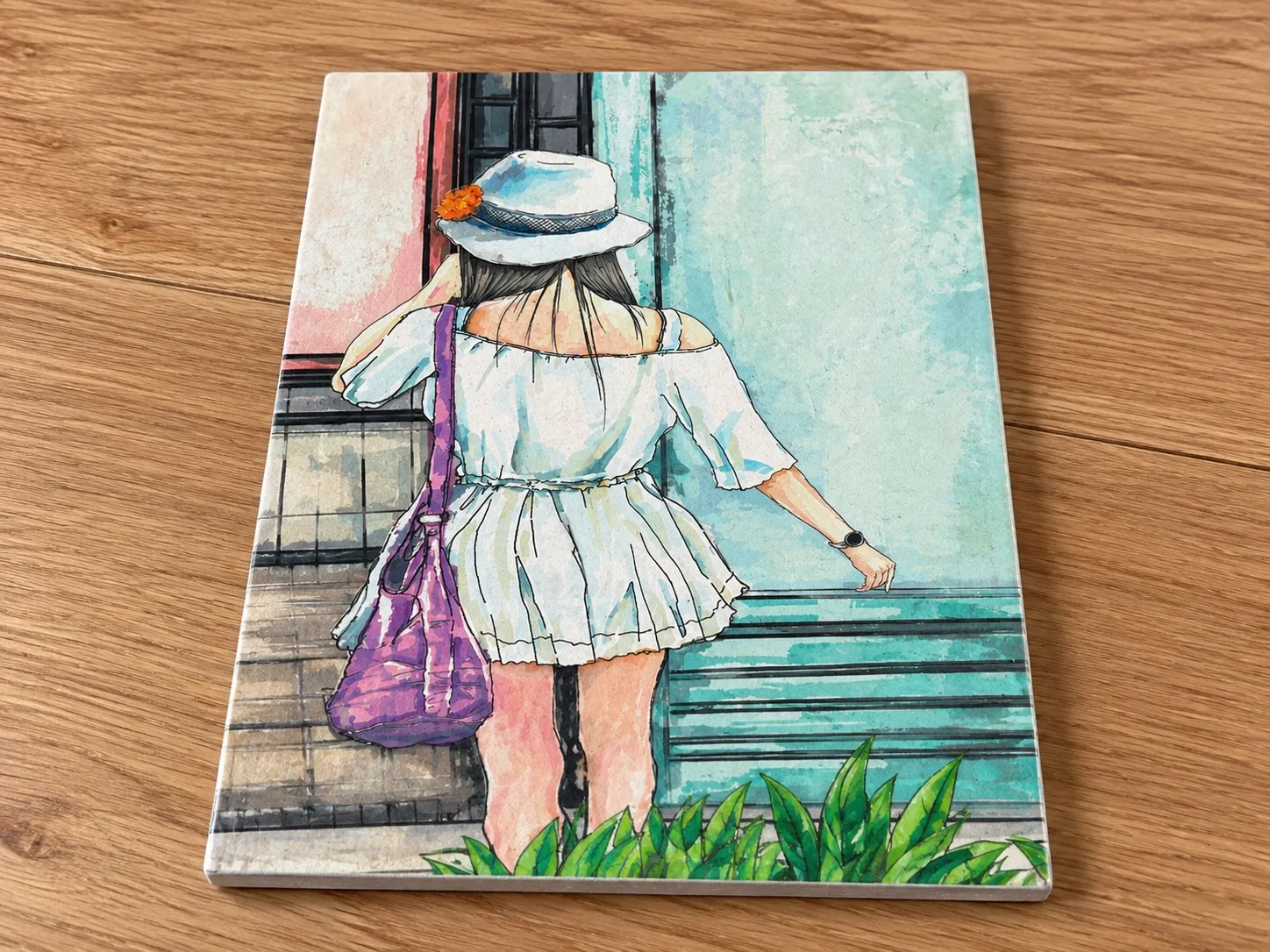 A rectangular unframed ceramic tile with an illustration of a woman in a white dress and hat, lying on a wooden table.