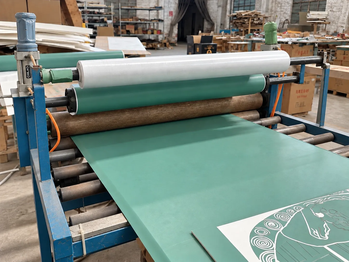 A roller laminator applying green resist film to a panel, with a cut and weeded mask on the panel in front.