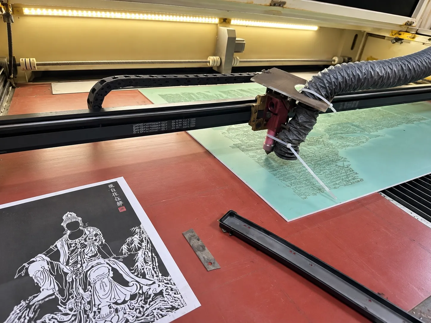 A laser head cutting a pattern into green resist film on a panel, a printed reference of the artwork on the bed beside it.