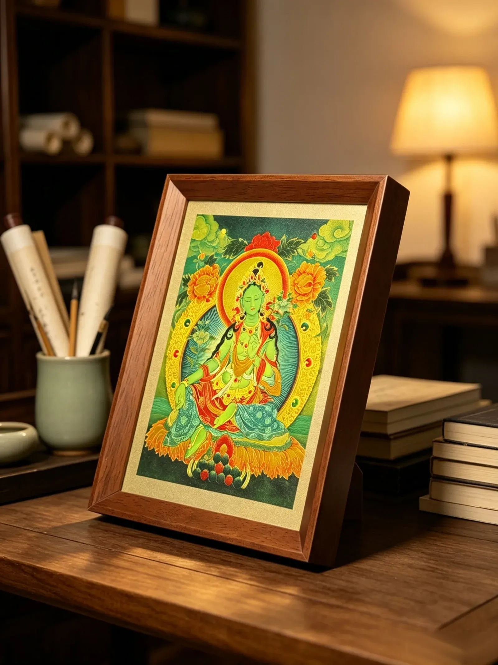 A Green Tara relief artwork in a wood frame standing on a study desk beside a brush pot, scrolls and a lamp.