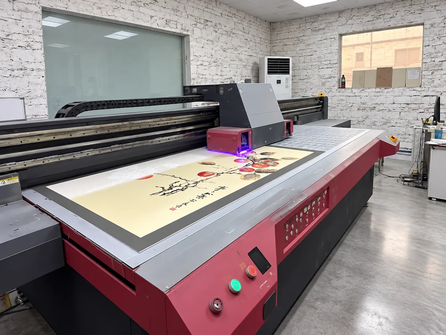 A UV flatbed printer mid-job, violet curing light glowing at the print carriage above a panel.